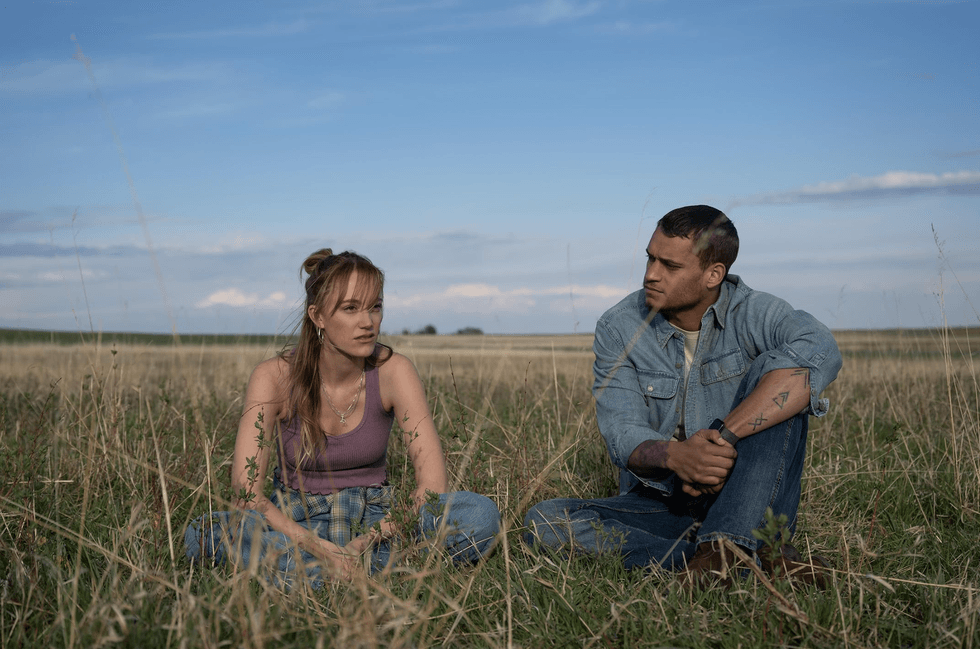 Two people sitting in a grassy field under a blue sky, engaged in conversation.