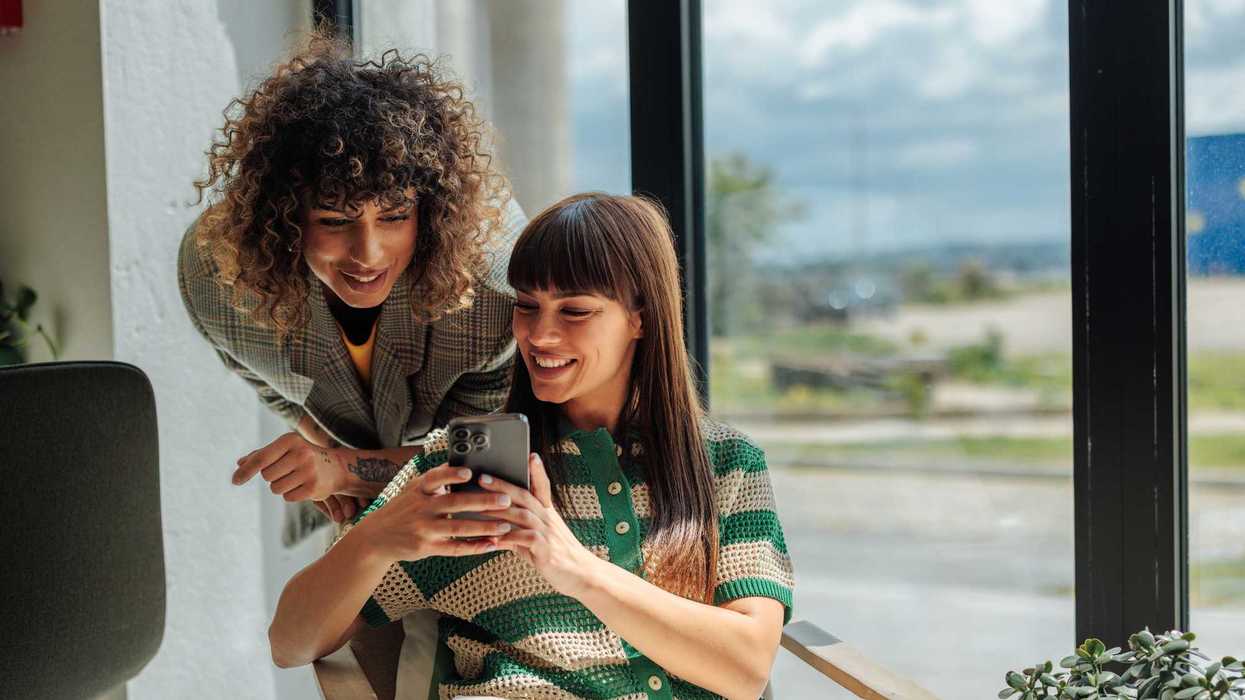 Two people smiling and looking at a smartphone indoors by a window.