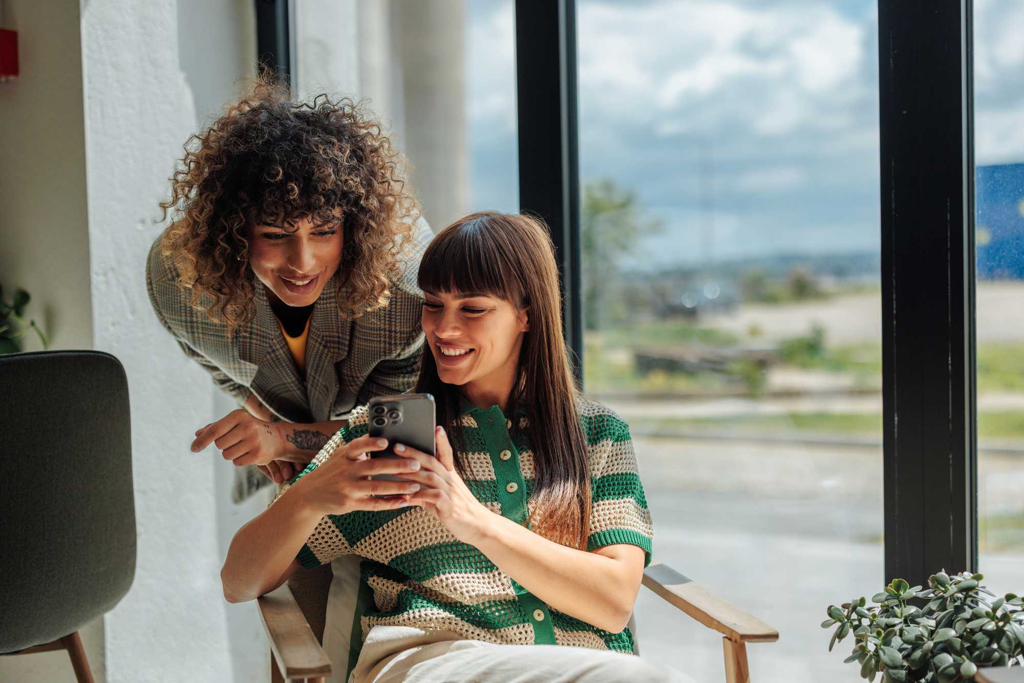 Two people smiling and looking at a smartphone indoors by a window.