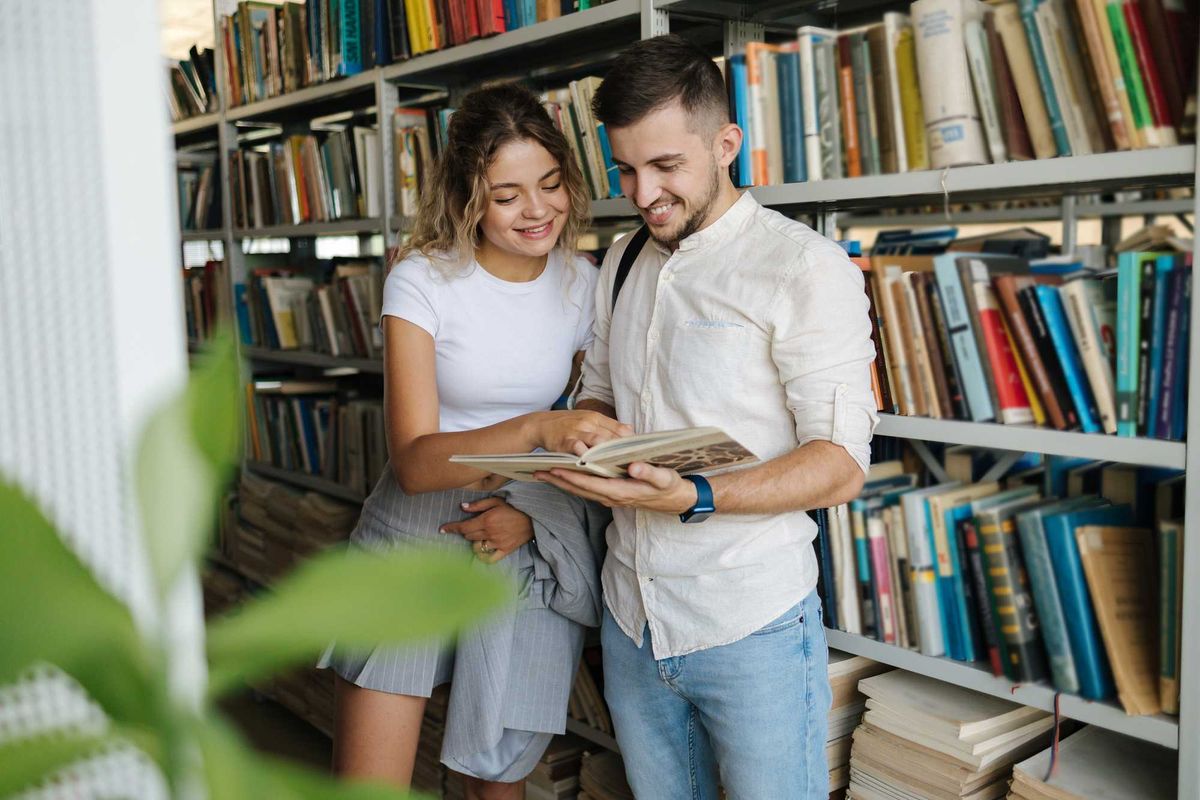 Two people smiling and reading a book in a library.