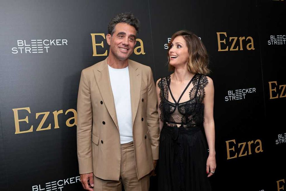 Two people smiling on the red carpet at an "Ezra" event.