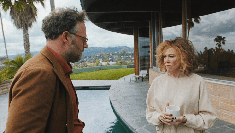 Two people stand by a pool, one holding a mug, with hills and clouds in the background.