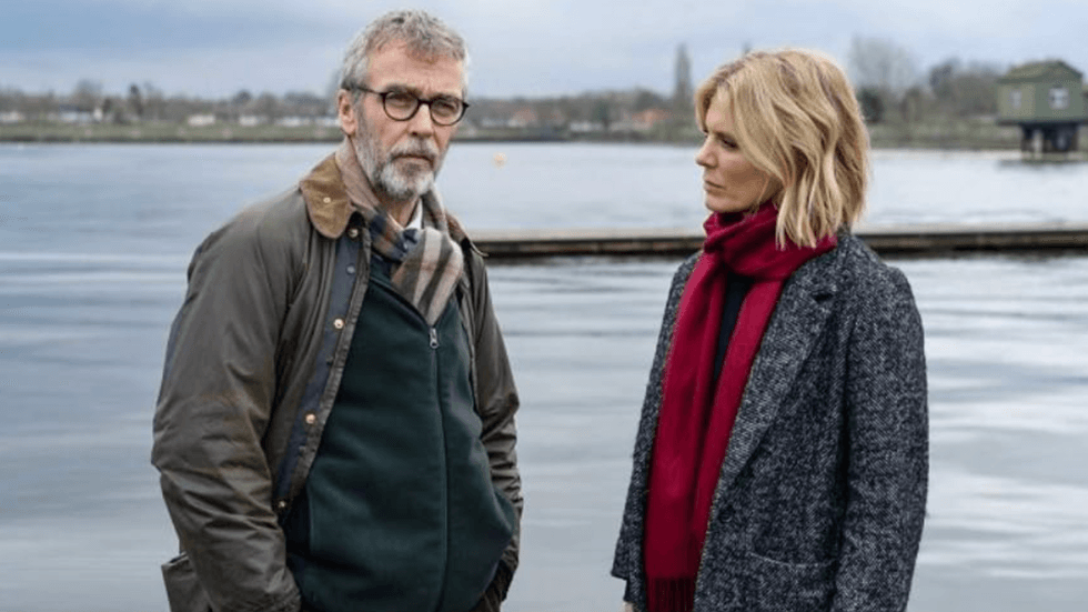 Two people stand by a serene lake, dressed warmly on an overcast day.