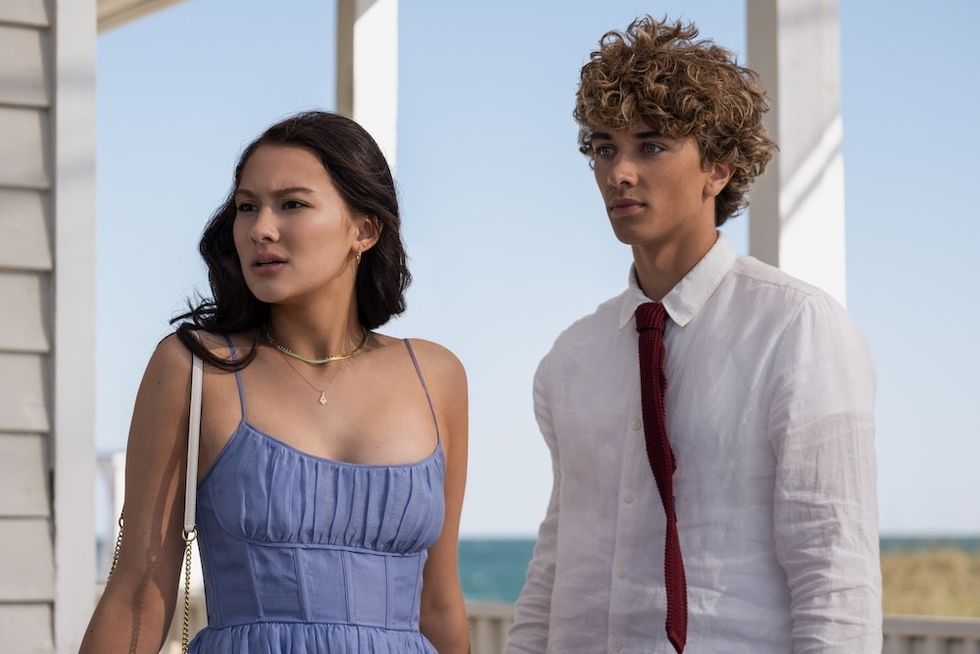 Two people standing on a porch by the sea, one in a blue dress, the other in a white shirt.