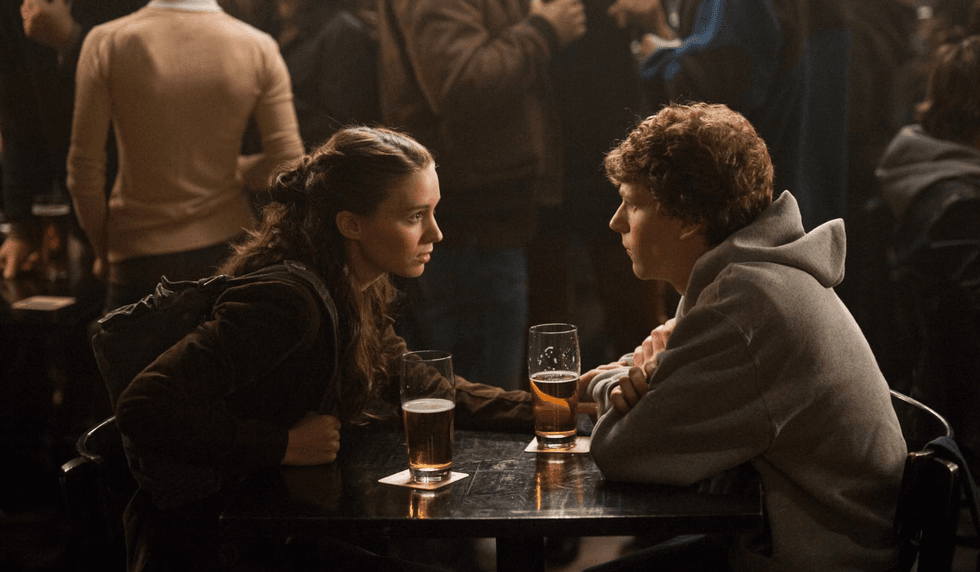 Two people talking at a bar table with drinks, surrounded by a lively crowd.