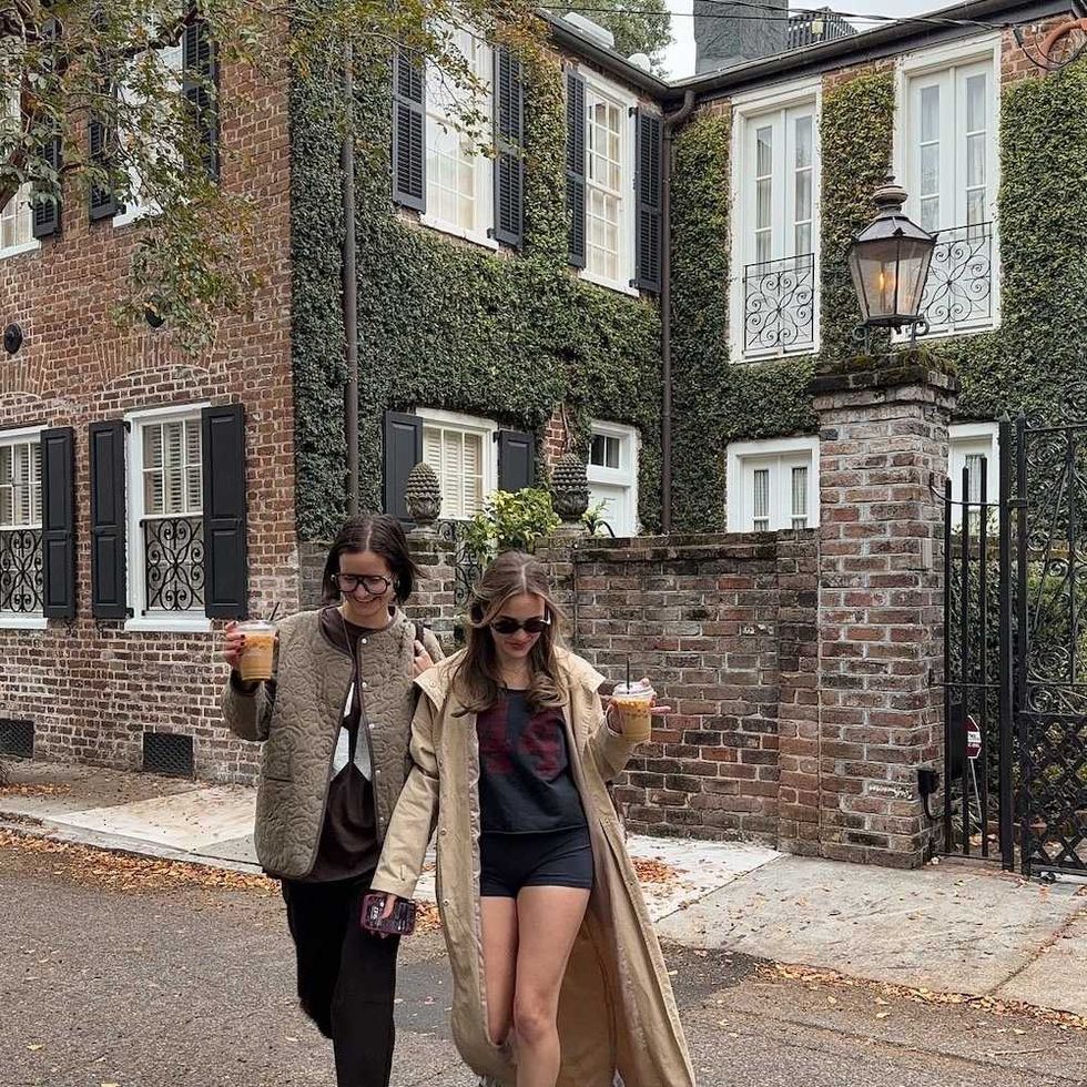 Two people walk with iced drinks in front of a brick house covered in ivy.