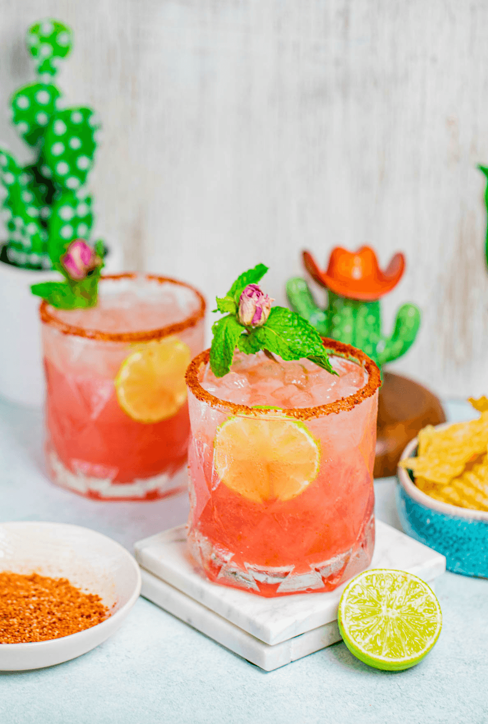 Two pink cocktails with lime slices, mint, and spicy rims next to lime and chips.
