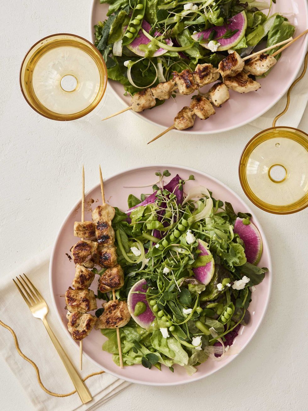 Two plates with salad and grilled chicken skewers, plus two golden-rimmed glasses.