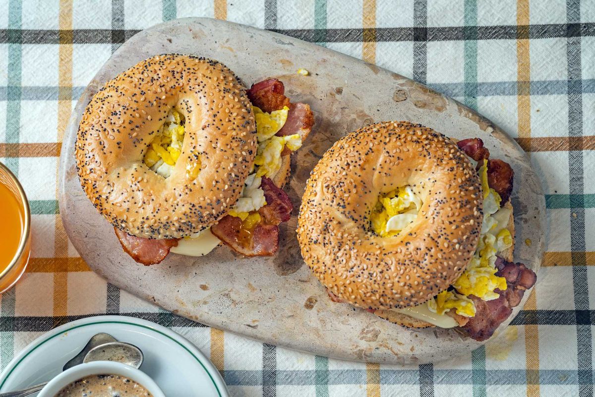 Two poppy seed bagel sandwiches with eggs and bacon on a stone board.