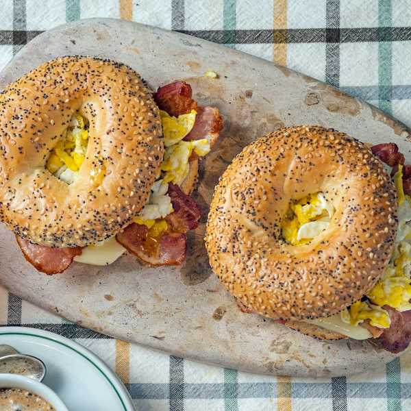 Two poppy seed bagel sandwiches with eggs and bacon on a stone board.