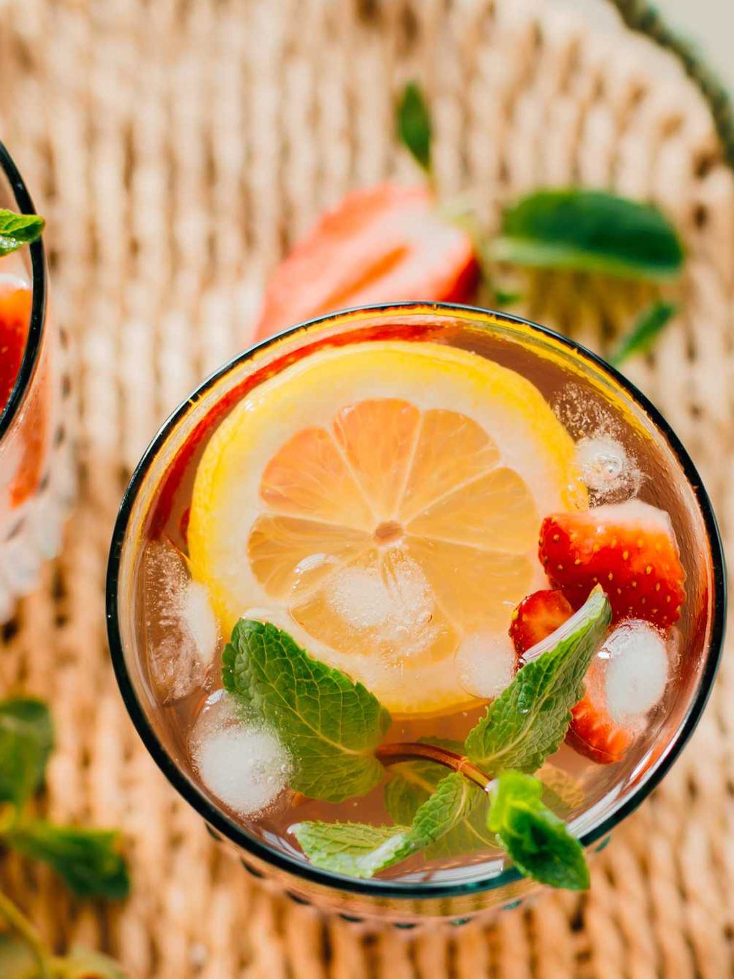 Two refreshing drinks with lemon, strawberries, mint, and ice on a woven mat.
