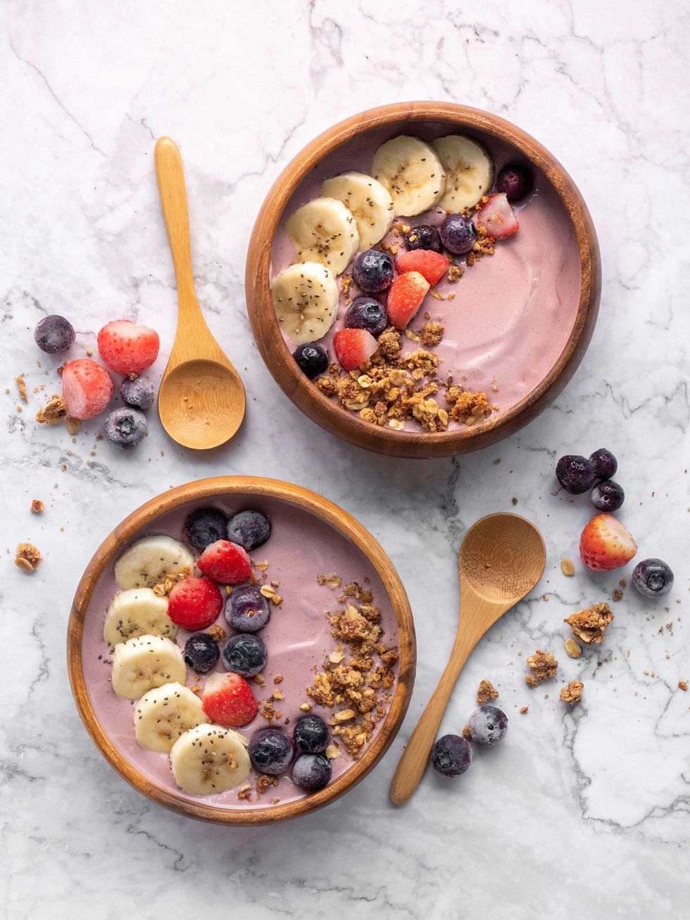 Two smoothie bowls with fruit, granola, and wooden spoons on a marble surface.