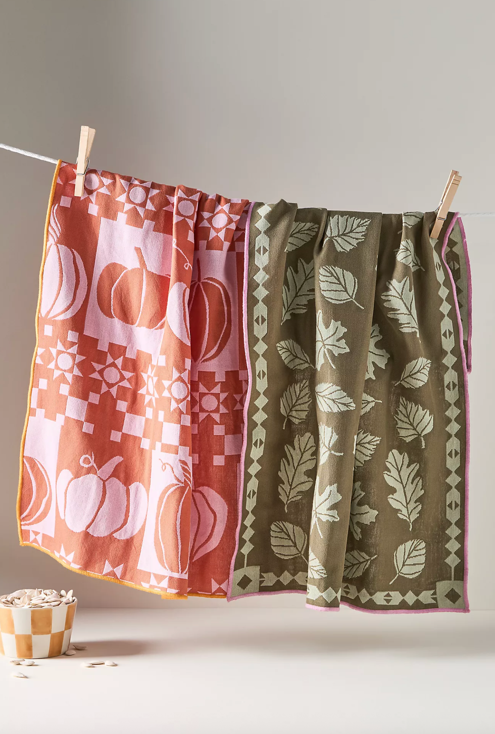 Two tea towels with pumpkin and leaf designs hanging on a line with clothespins.