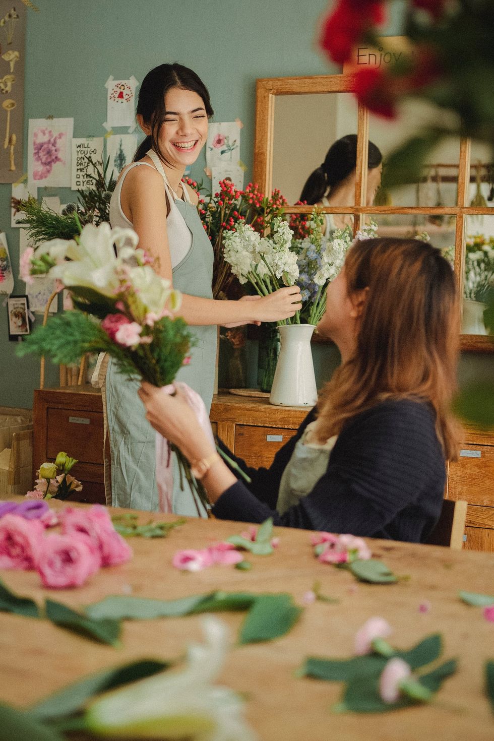 Two women arranging flowers and smiling in a cozy, decorated room.