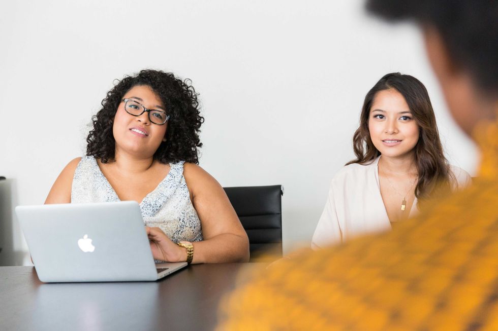 Two women at a meeting table, one with a laptop, facing a blurred person.