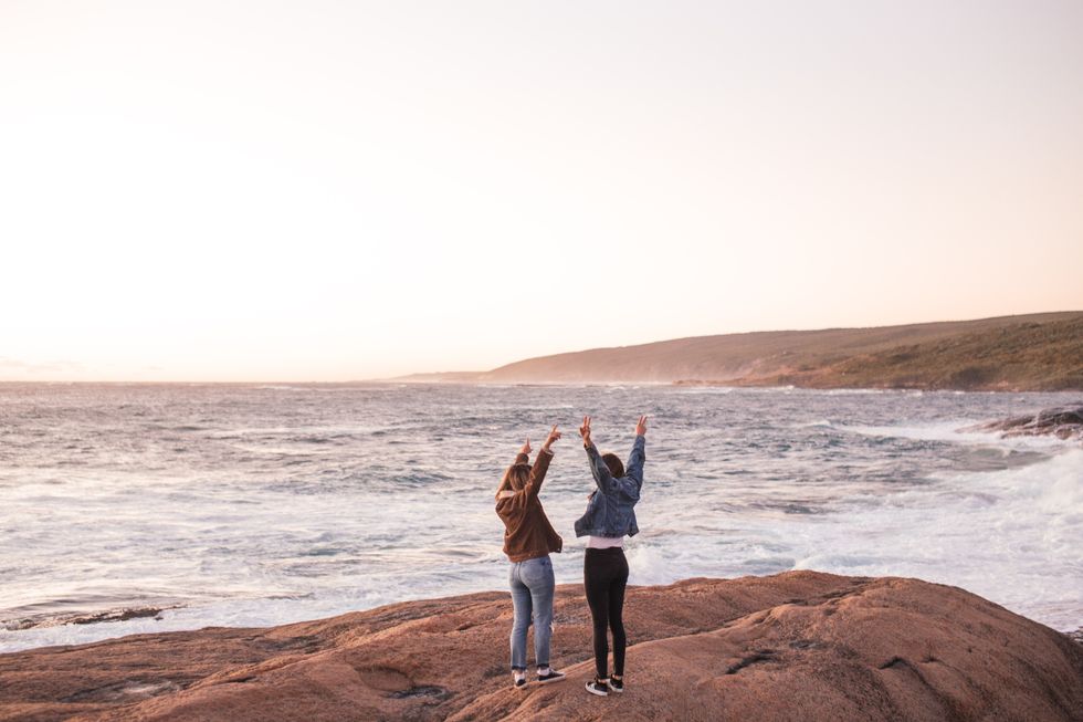 two women by the sea standing on a rock with arms up in excitement