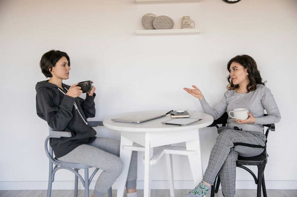 Two women chat over coffee at a white table, one gesturing, the other listening.