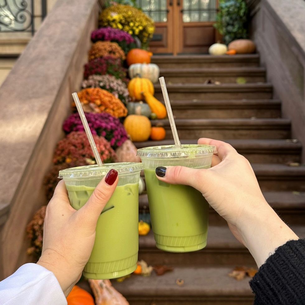 two women cheersing iced matcha lattes in front of a stoop decorated for fall