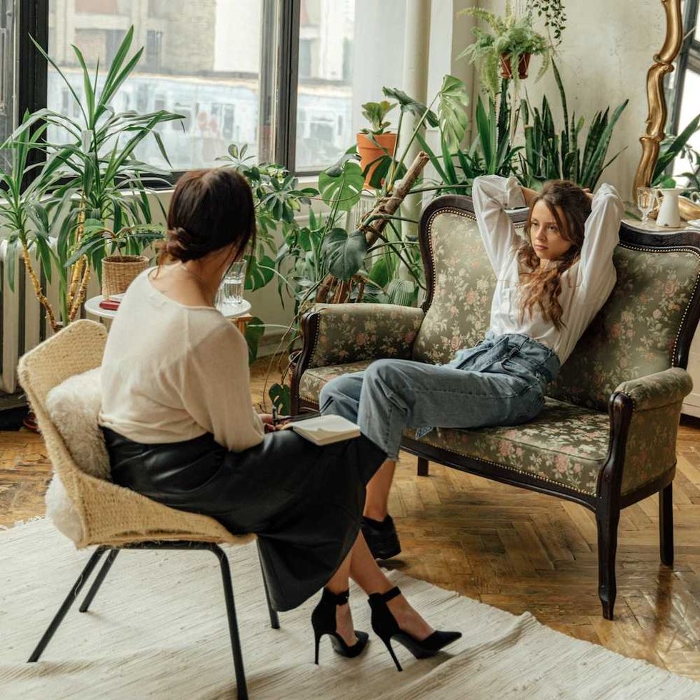 Two women conversing in a plant-filled room, one writing in a notebook.