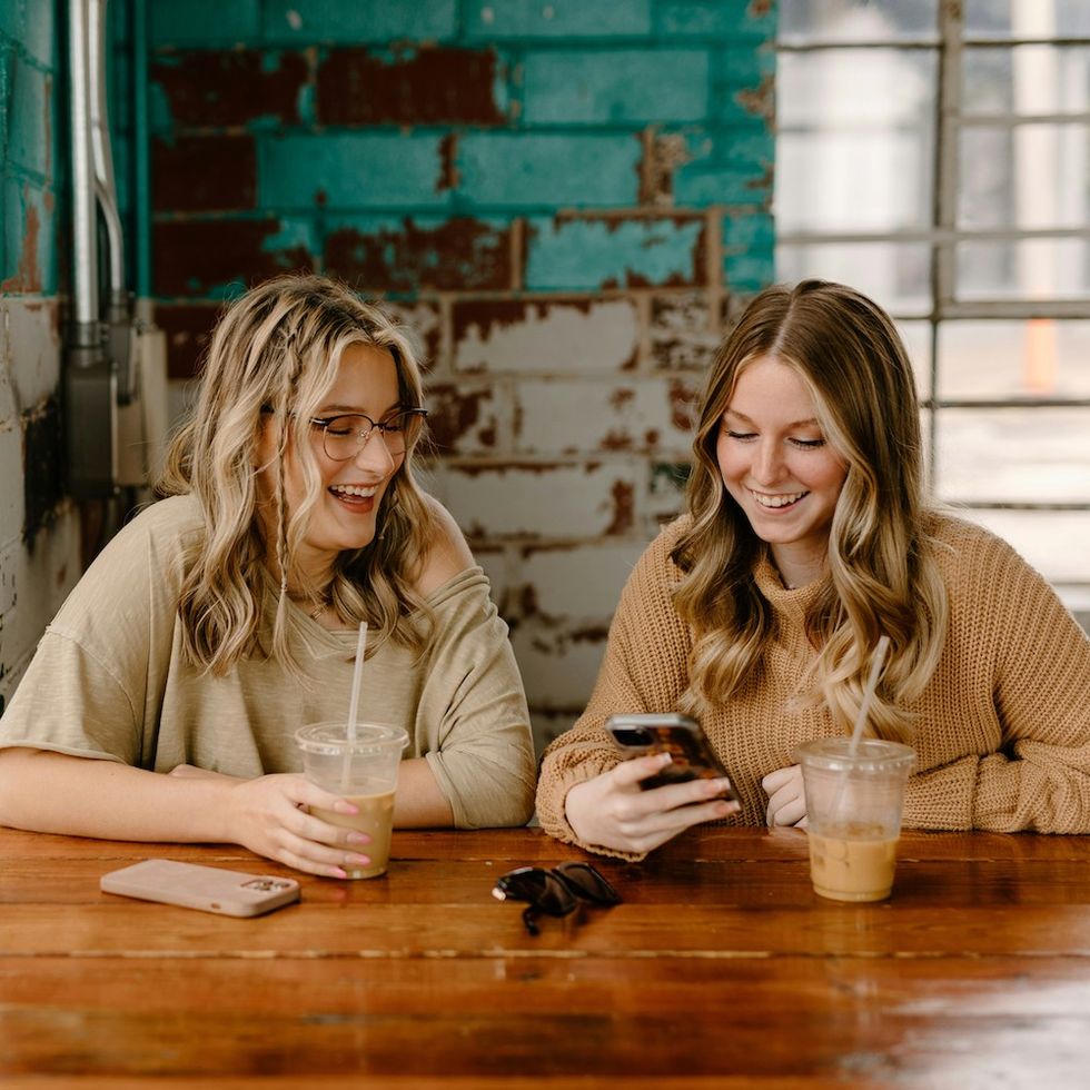 two women drinking iced coffee at a table together, looking at an iPhone