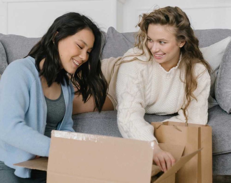 Two women happily unpack items from cardboard boxes on a couch.