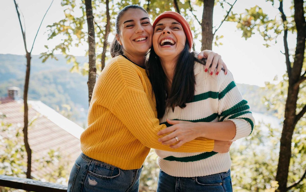 Two women hugging and laughing outdoors on a sunny day.