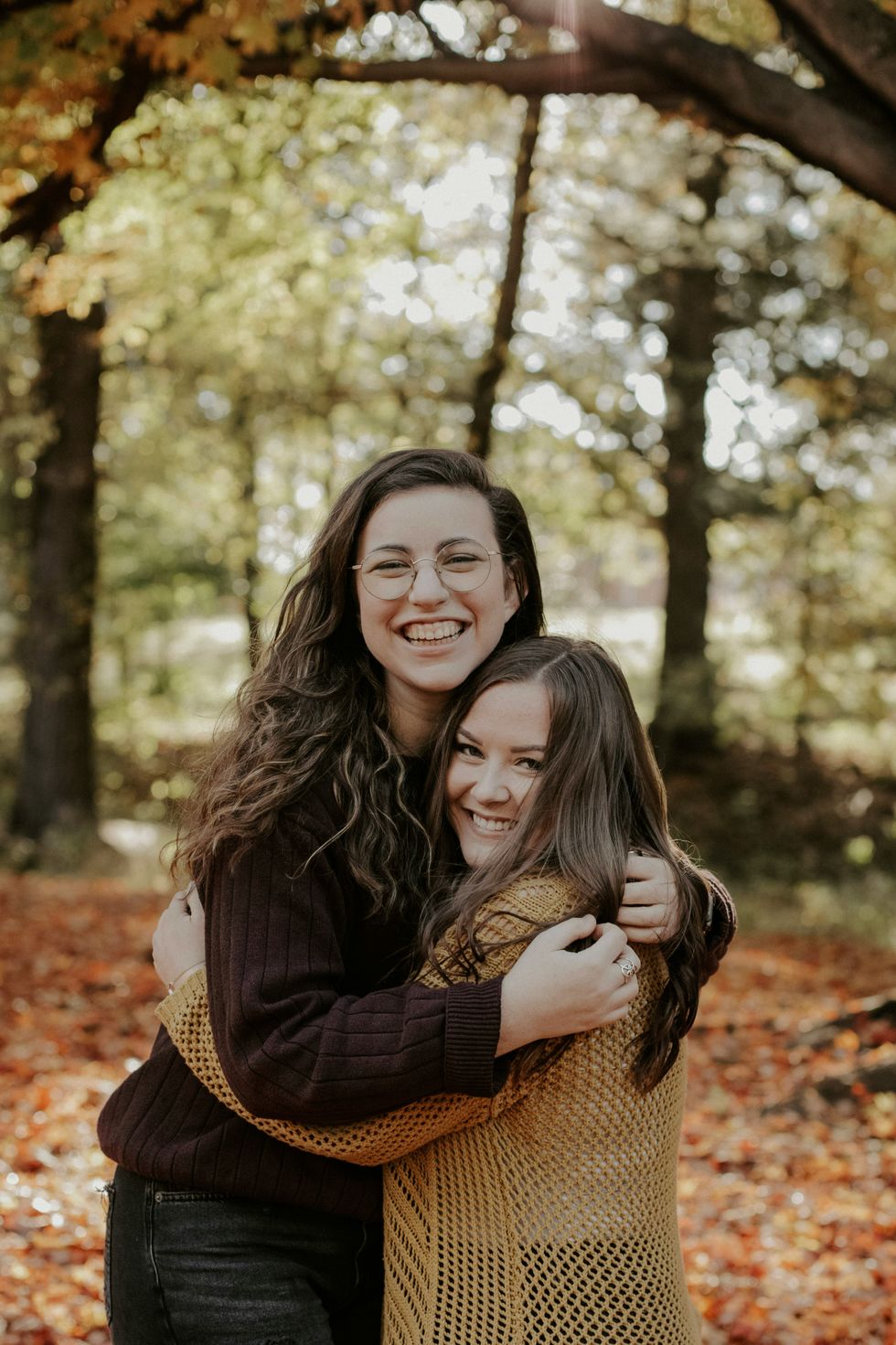 Two women hugging and smiling in a sunlit forest with autumn leaves.