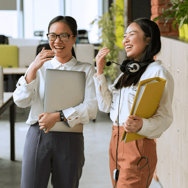 Two women in an office laughing, holding a laptop and files, with one wearing a headset.