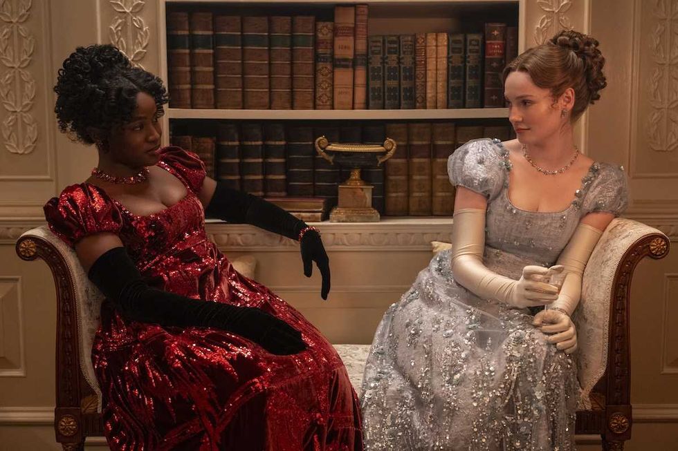 Two women in elegant dresses sit on a couch, surrounded by bookshelves. bridgerton season 4 episode 5