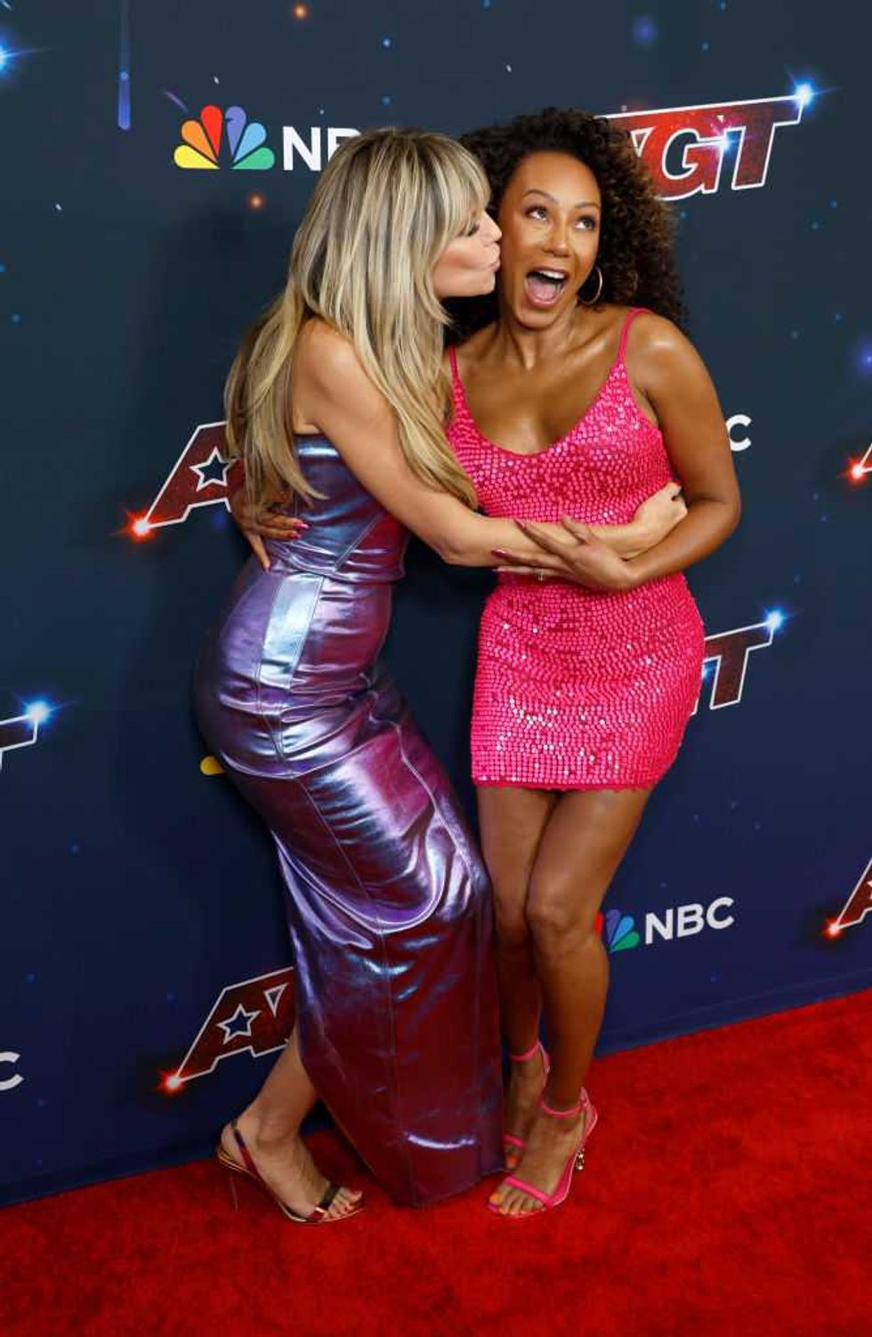 Two women in shiny dresses embrace and smile on a red carpet.