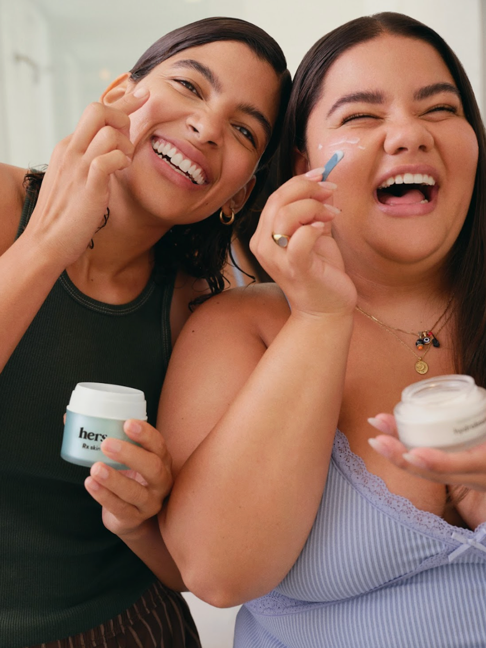 Two women joyfully applying skincare cream together.