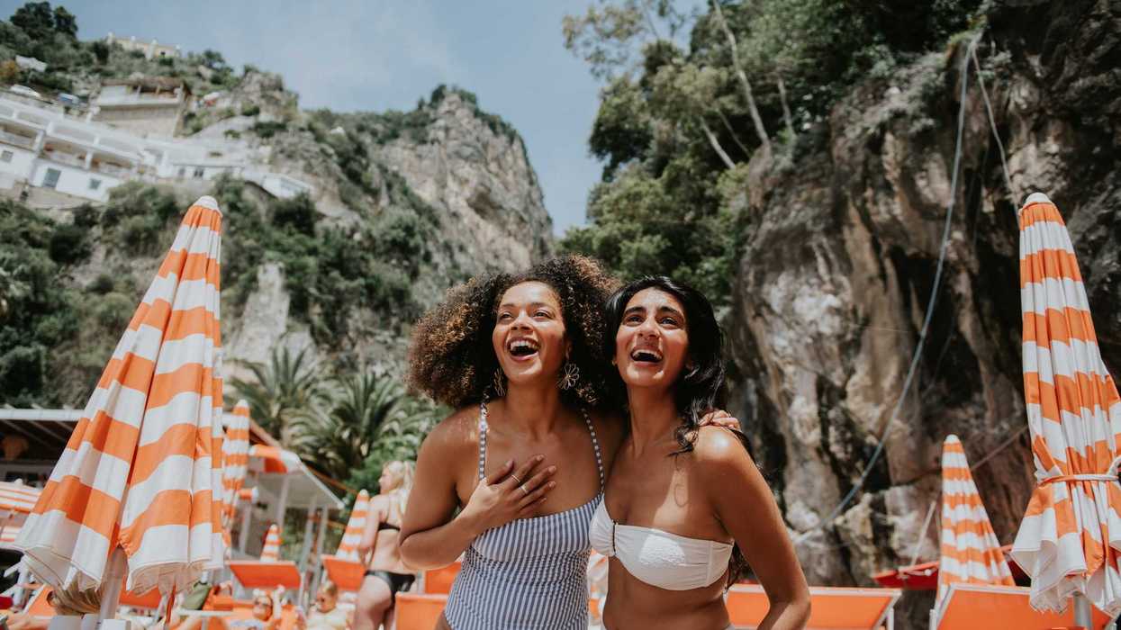 Two women laughing at a beach with striped umbrellas and rocky cliffs.