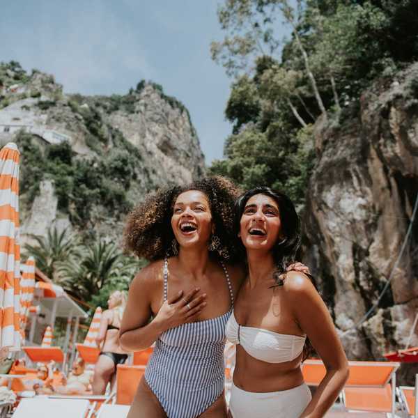 Two women laughing at a beach with striped umbrellas and rocky cliffs.