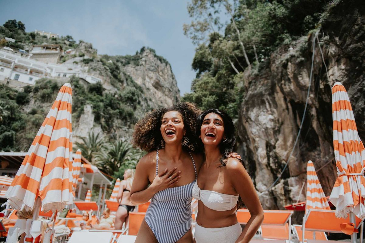 Two women laughing at a sunny beach with striped umbrellas.