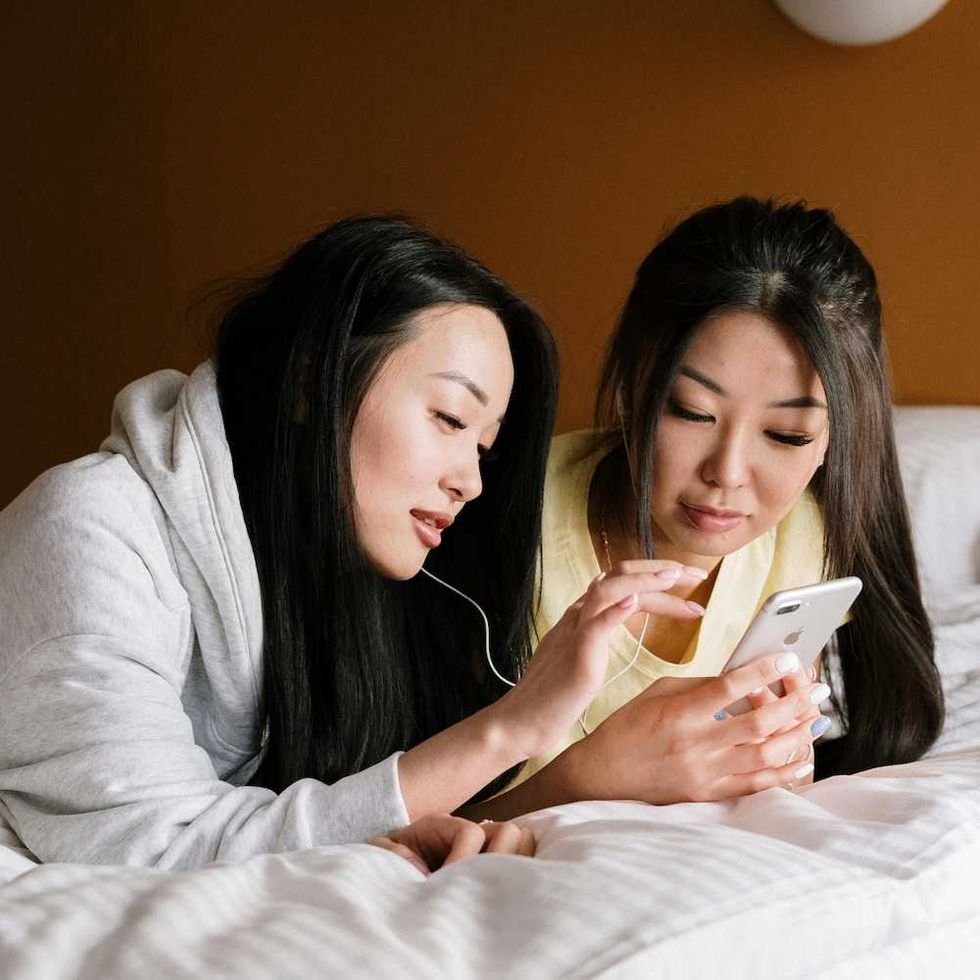 Two women lying on a bed, sharing earphones and looking at a smartphone.