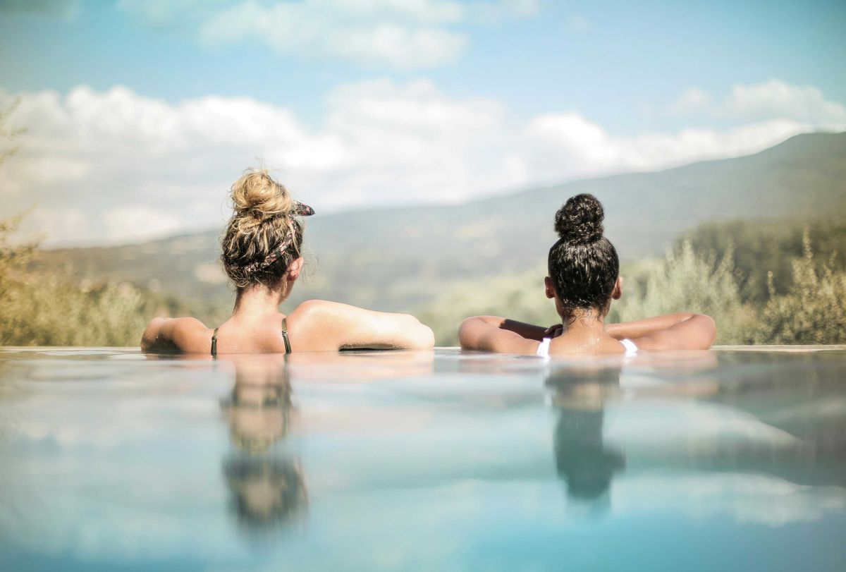 Two women relax in an infinity pool facing scenic mountain views under a blue sky.