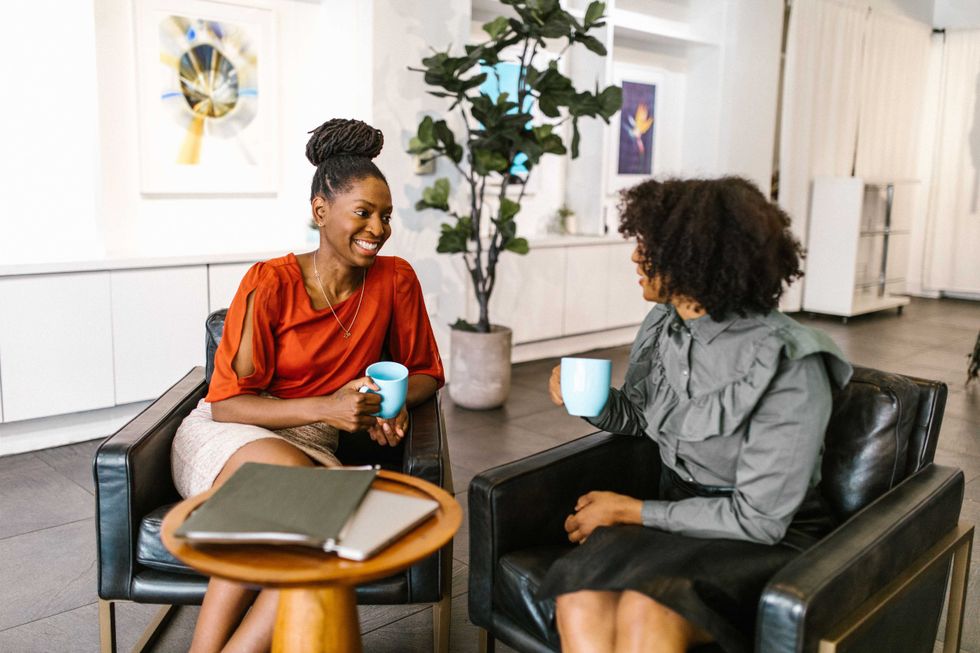 Two women sit and chat over coffee cups in a cozy, modern office setting.