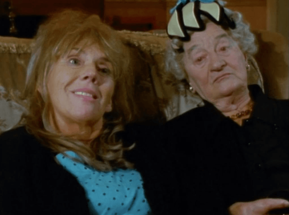 Two women sitting on a couch, one smiling, the other wearing a hat with a neutral expression.