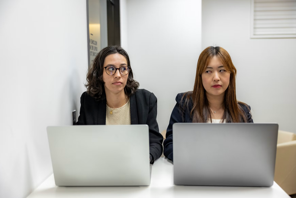 Two women sitting with laptops, exchanging doubtful glances in an office.