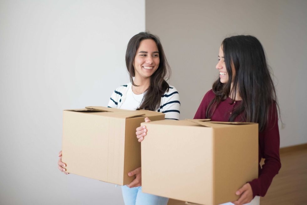 Two women smiling and carrying cardboard boxes in a room.