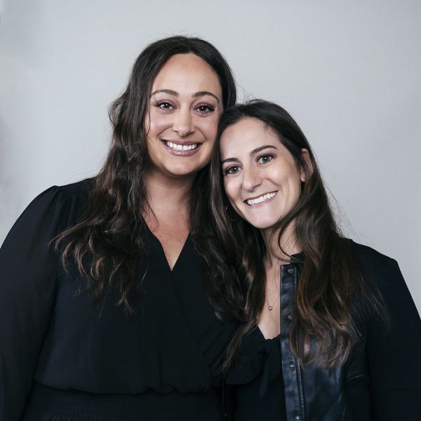 Two women smiling and standing closely together against a plain background.