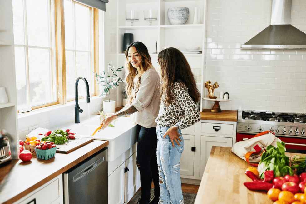 Two women smiling and washing vegetables in a bright kitchen.
