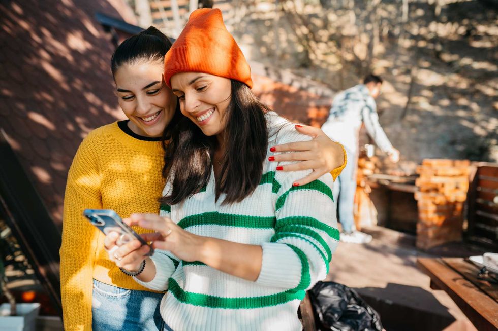 Two women smiling at a phone outside; one wears a beanie.