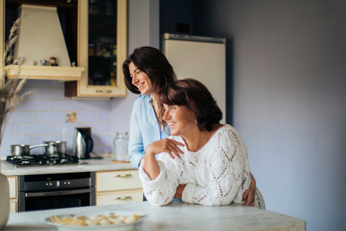 Two women smiling in a kitchen, leaning on a counter with food.