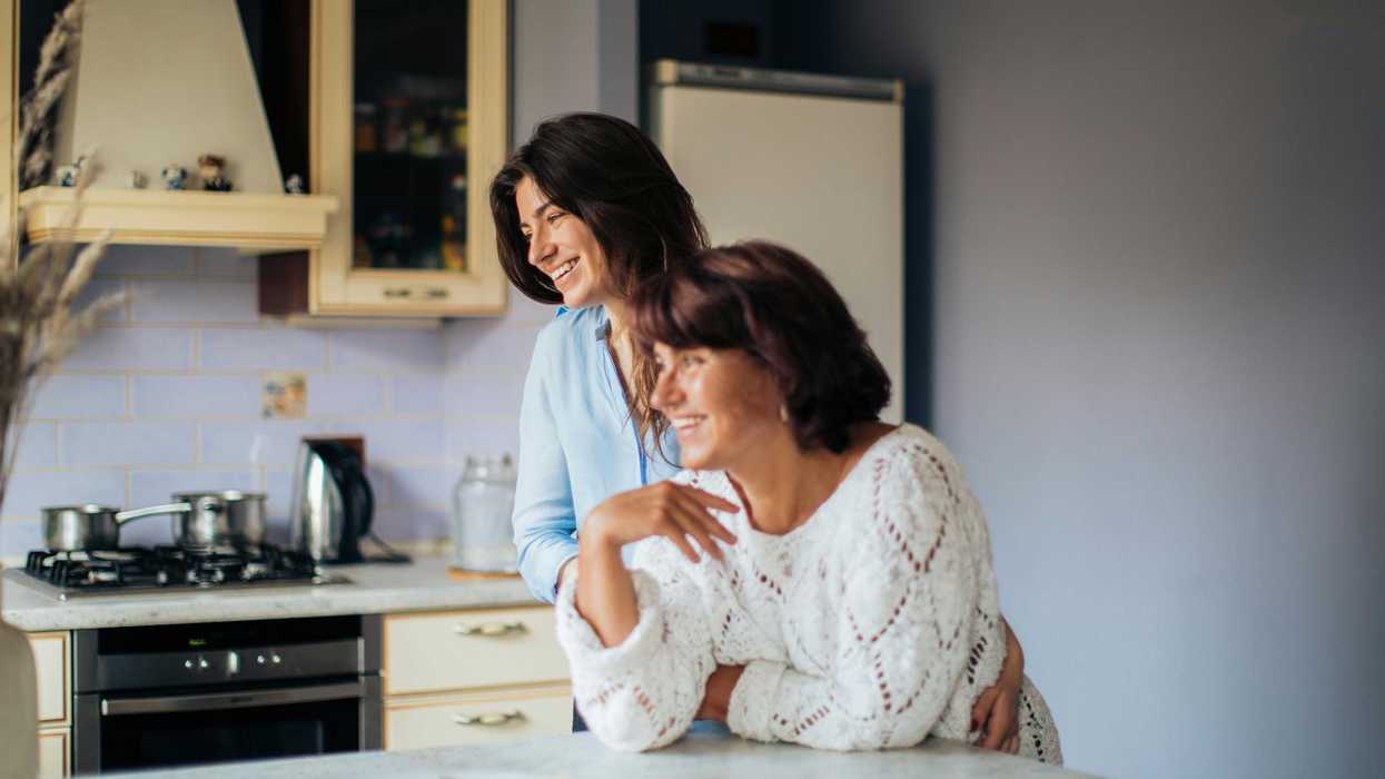 Two women smiling in a kitchen, leaning on a counter with food.