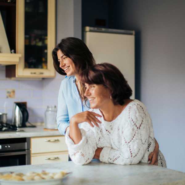 Two women smiling in a kitchen, leaning on a counter with food.
