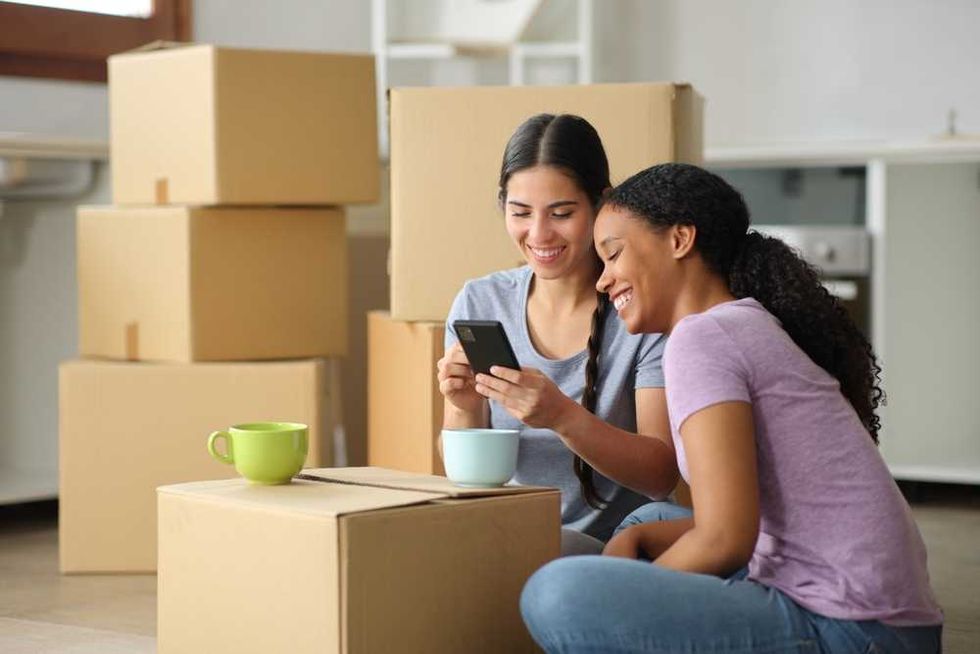 Two women smiling, looking at a phone, surrounded by moving boxes.