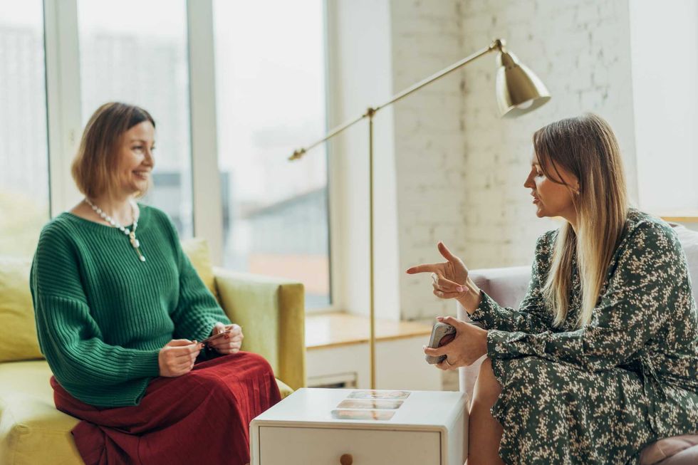 Two women talking in a cozy room with a lamp and large window.