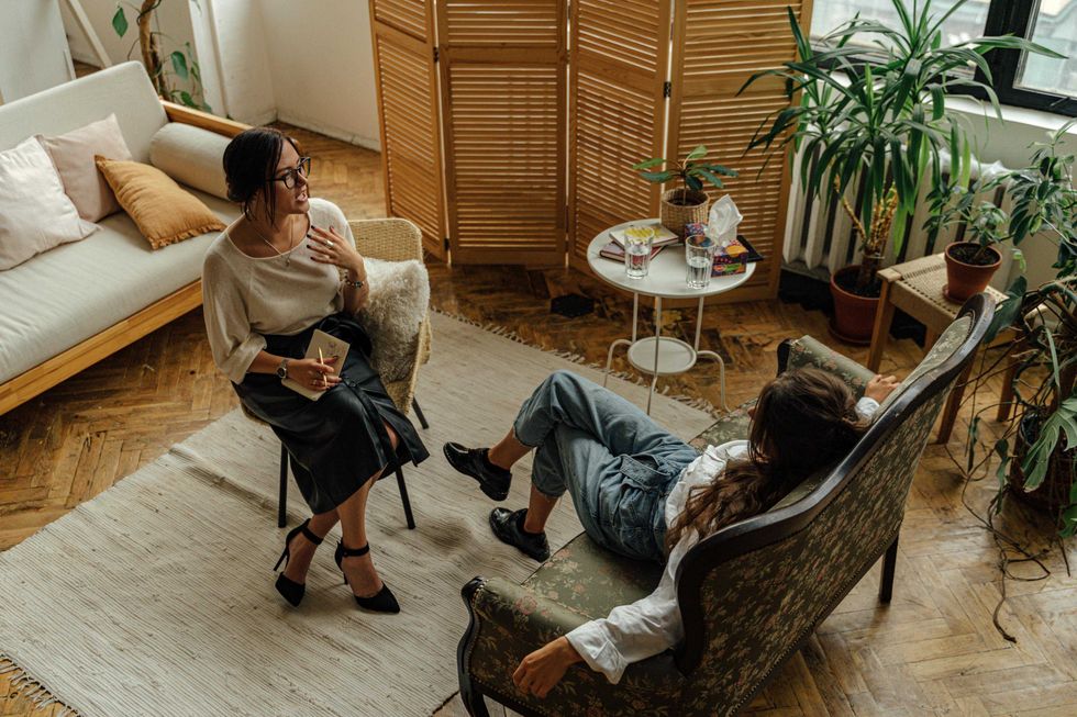 Two women talking in a cozy room with plants and wooden decor.