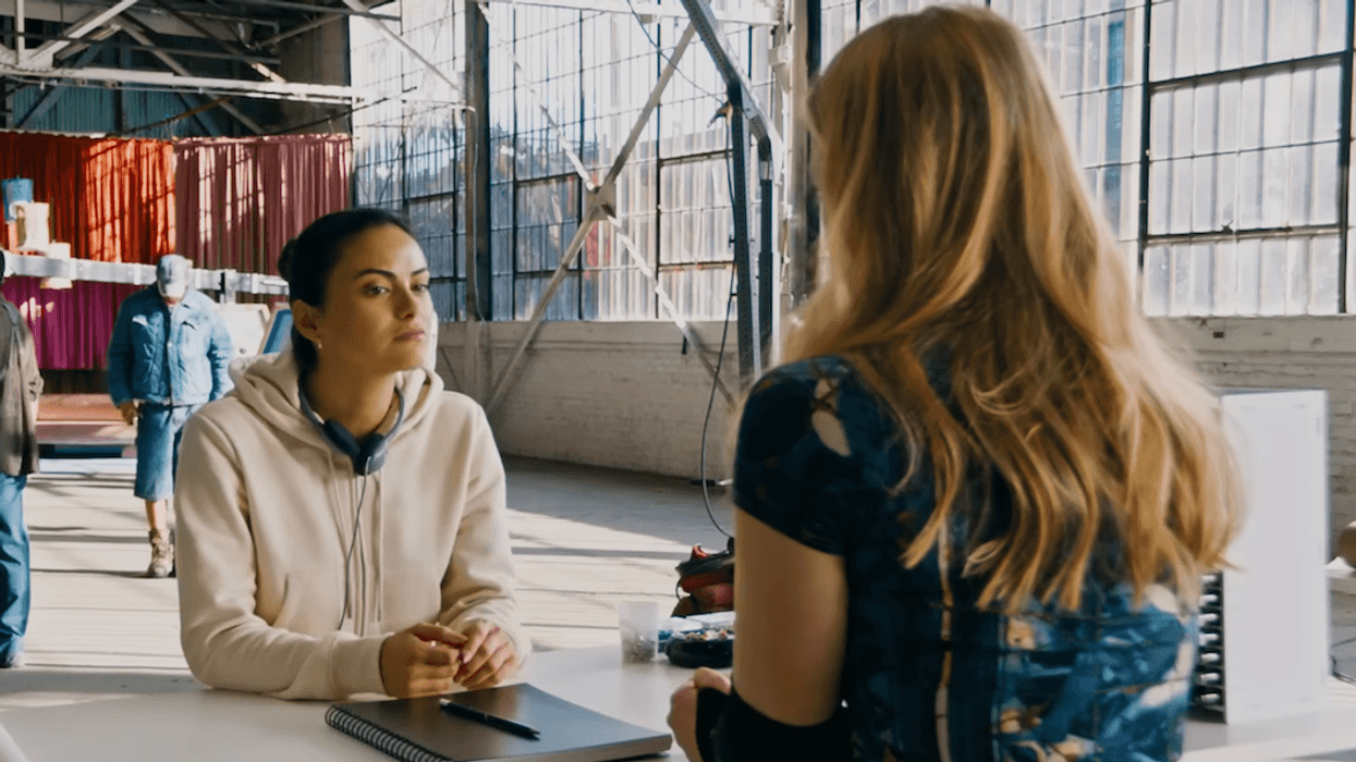 Two women talking in a sunlit warehouse with people and equipment in the background.